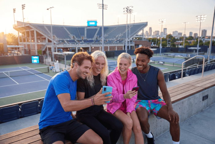 Friends looking at smartphones together in a modern setting, laughing and engaged, with tennis court or stadium visible in background